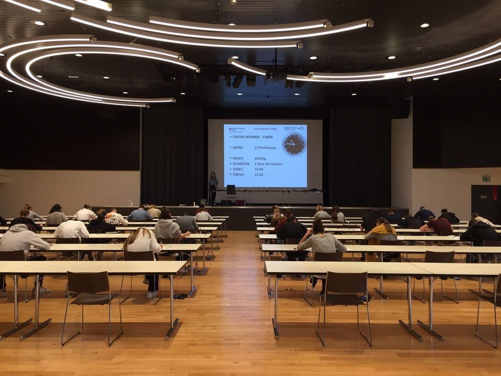 A big exam hall filled with long chairs. Students sit on the tables to write their exam. In the front is a presentation shown with a clock and some exam information.