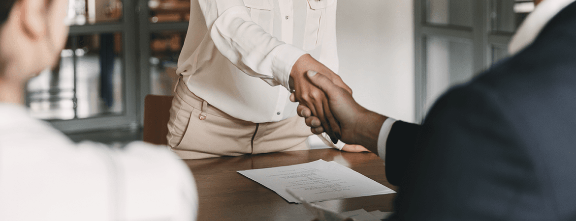 An individual person in a job interview. Papers like the CV and applicaiton letter lay on the table. The job candidate and the employer are shaking hands.