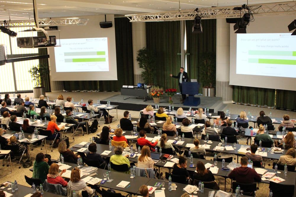 Many teachers sitting in a plenary hall listening to the presenter. Two big screens show the slides of the plenary speakers' presentation. The speaker is standing on a stage. The teachers all have papermaterial or laptops in front of them to take notes or
