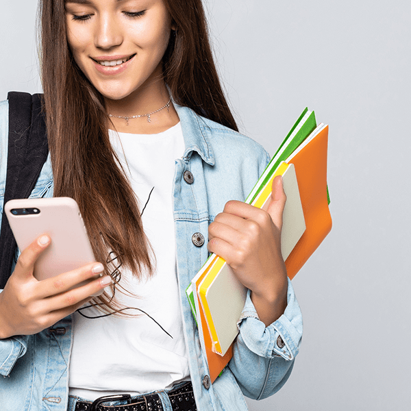 A girl holding a phone in her right and some school documents for her Cambridge English Exam Preparation in her left hand. She is looking into the phone where she uses Test & Train by Cambridge for digital Exam Preparation.