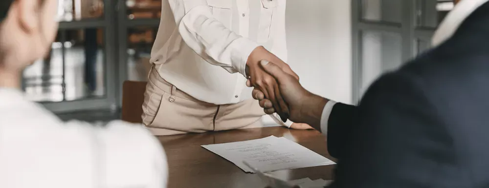 An individual person in a job interview. Papers like the CV and applicaiton letter lay on the table. The job candidate and the employer are shaking hands.