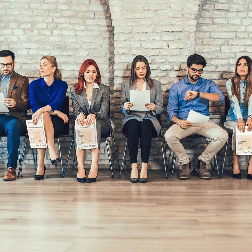 Six people, four women and two male, are sitting on chairs waiting to get picked up for their job interview. They are all holding their certificates and job interview documents in their hands.